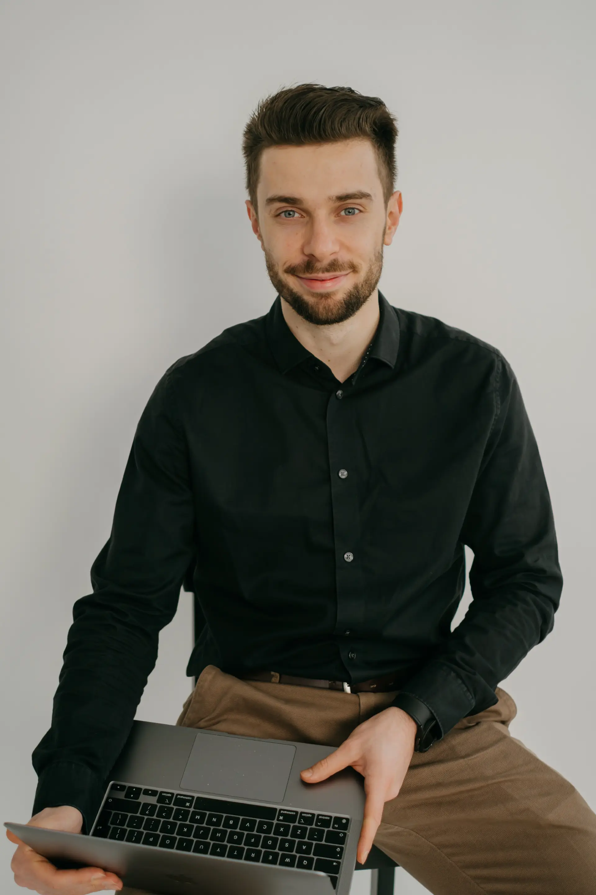 Profesjonalny portret Jakuba Budziocha, współzałożyciela agencji WaveMed, ubranego w czarną koszulę i brązowe spodnie. Trzyma laptopa i uśmiecha się pewnie, siedząc na prostym białym tle.